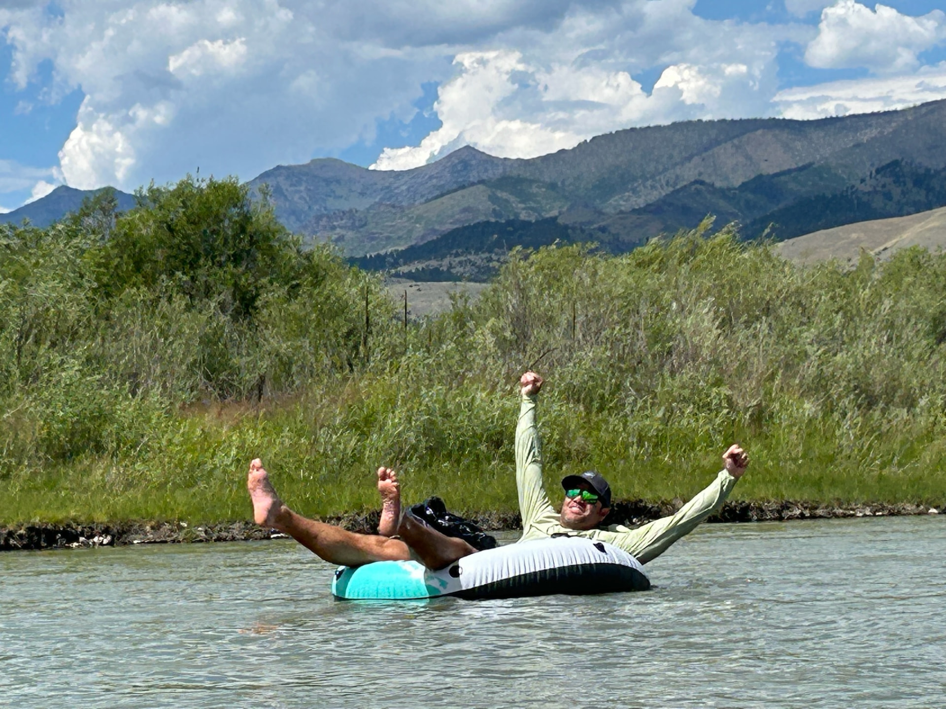 Arms raised, floating down a river on a tube with mountains behind
