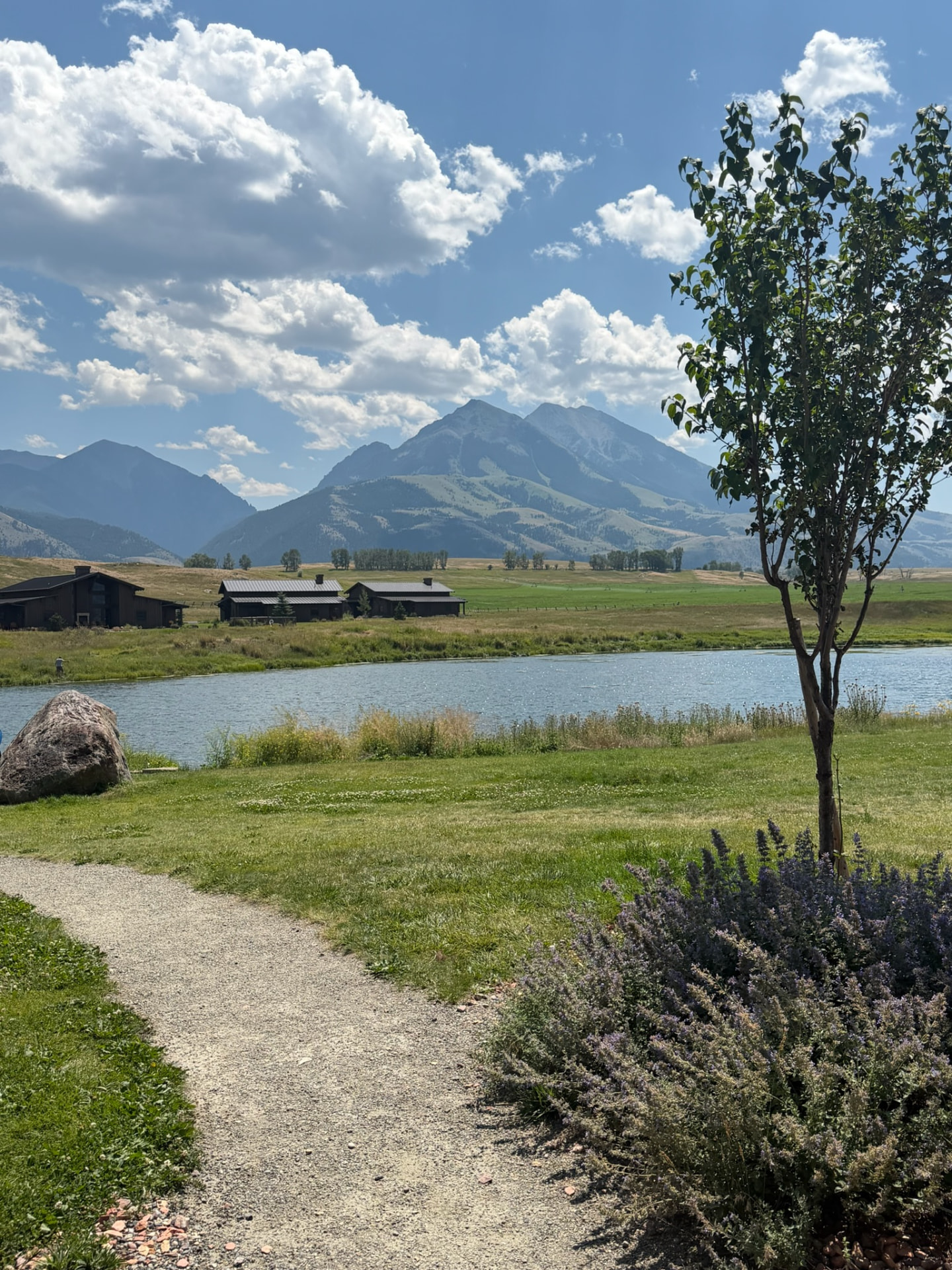 A gravel path leading to a pond with dark ranch barns and mountains beyond